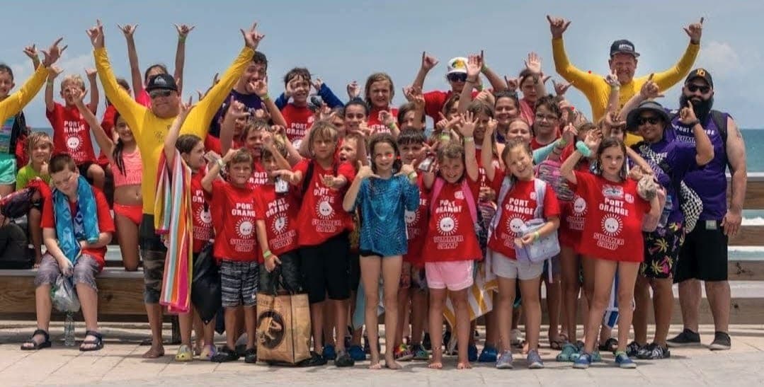 Big group of summer camp kids and instructors throwing shakas together on the boardwalk at Daytona Beach, ocean behind them
